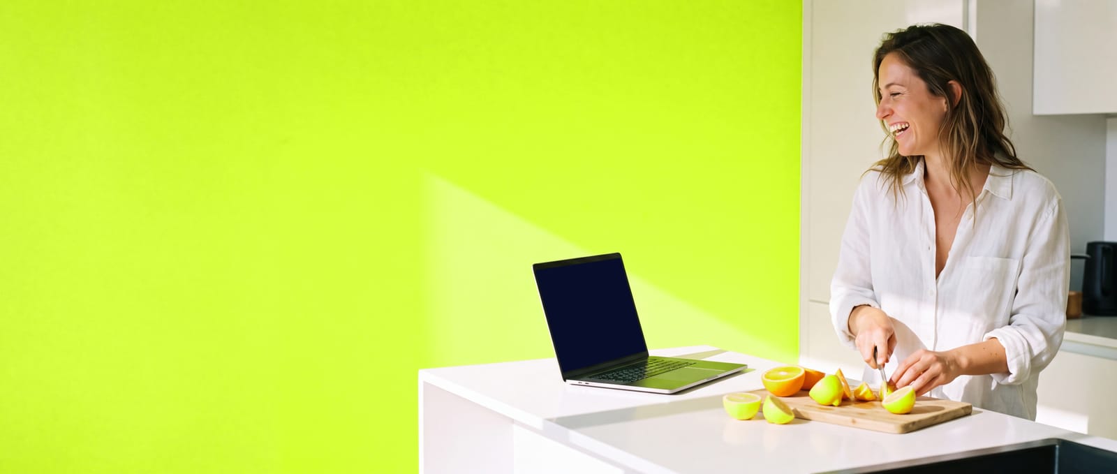 A woman laughing as she slices citrus at a white kitchen island, her laptop open beside her against a volt wall.