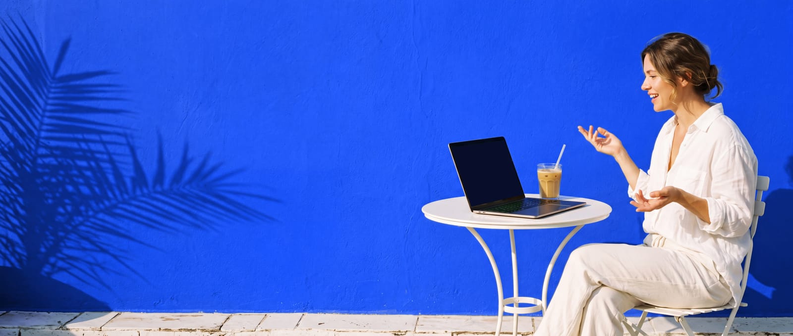 A woman in white linen at a café table on a sunny terrace, talking to her laptop against a cobalt wall.